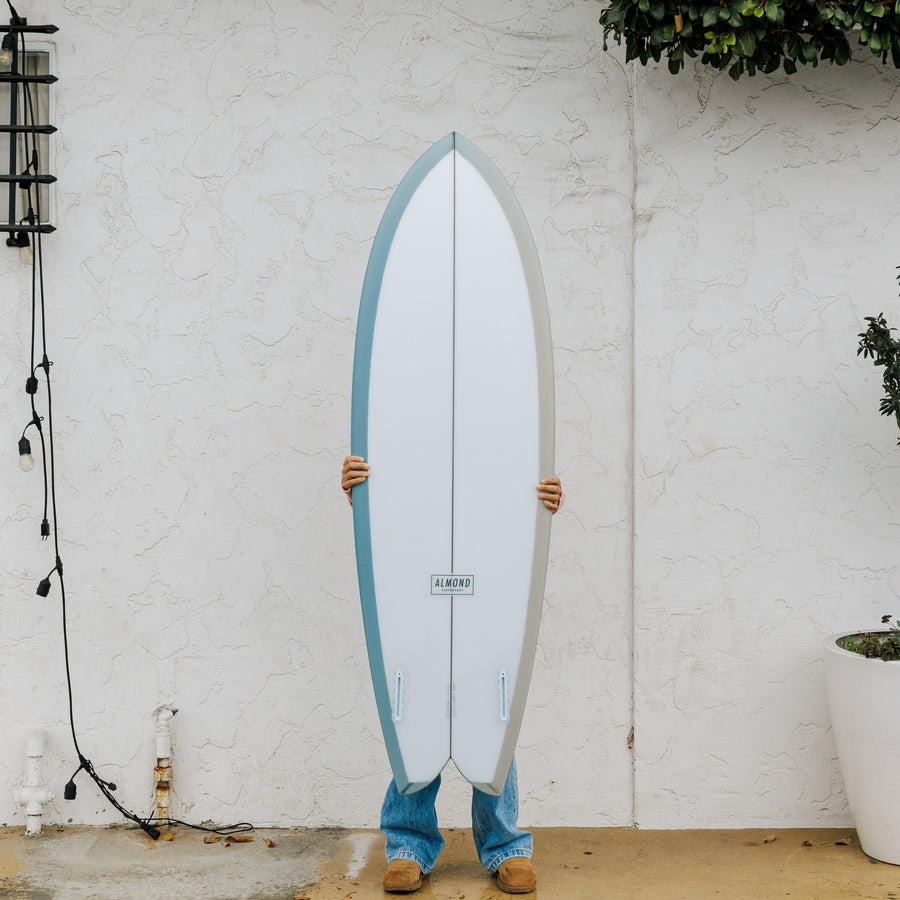 Person holding a surfboard with a white and blue design against a white brick wall.