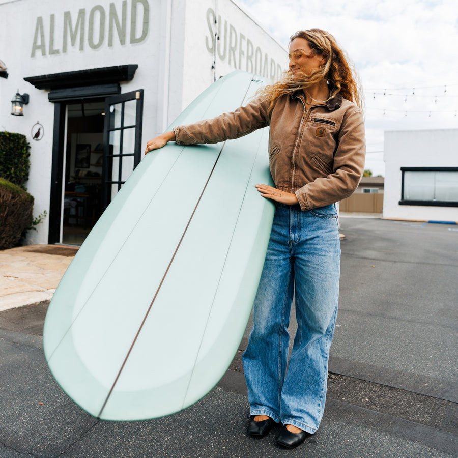 Person holding a surfboard in front of a building with 'Almond Surfboards' on it.